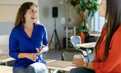 A woman wearing blue top in focus talking to another woman facing away from the camera.