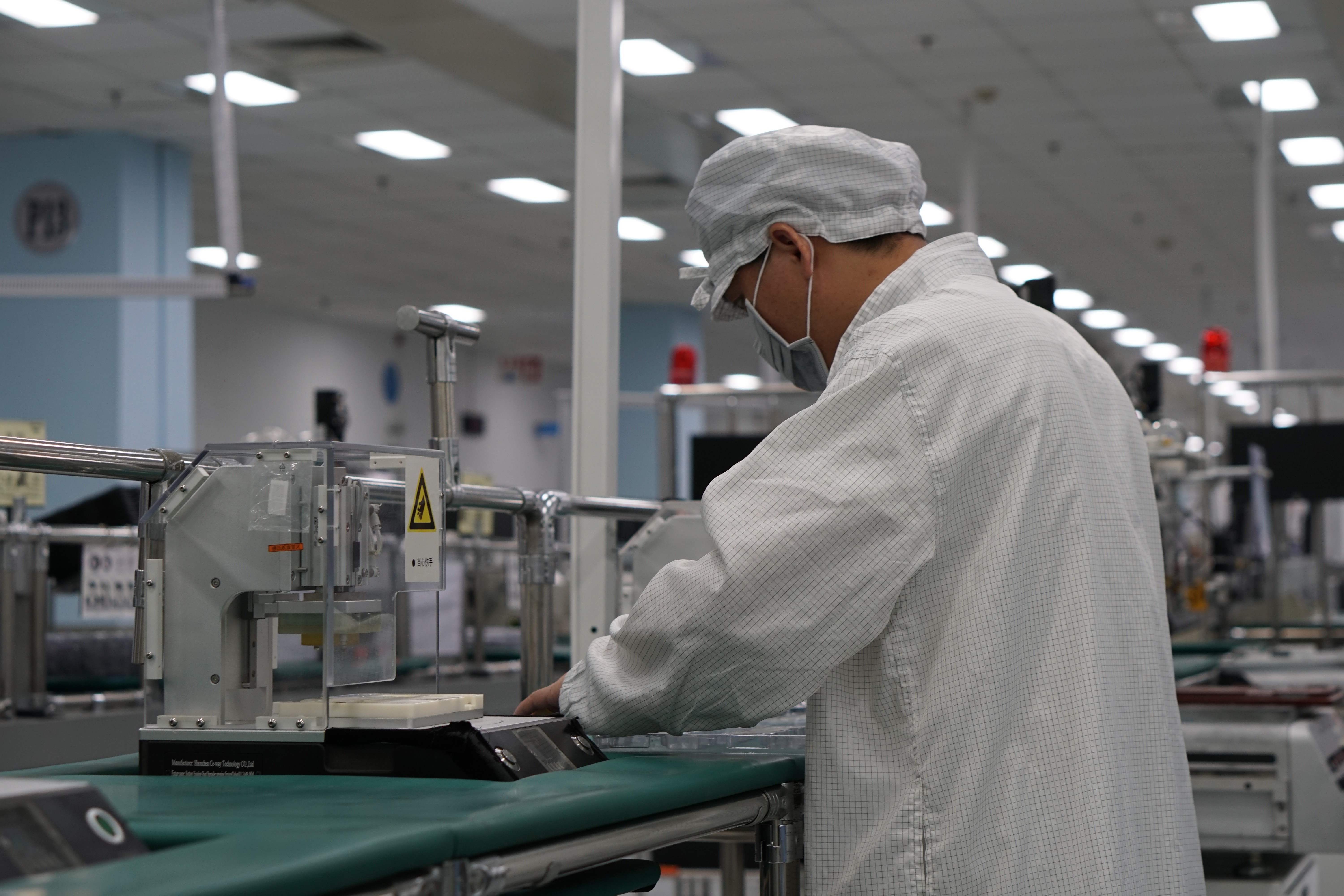 A factory worker concentrating on a task at their station, wearing a checkered factory uniform, cap and mask.