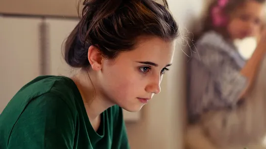 Focused teenage girl writing in a notebook while working on a laptop at a desk