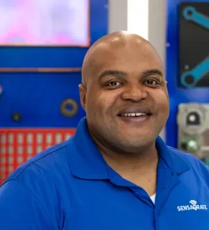A smiling man wearing a bright blue polo shirt with the Sensagrate logo on the chest, positioned in front of a blue background.