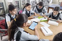 Four female students in school uniforms sit around a wooden table in a library, collaborating while using Samsung tablets and unboxing educational tech kits.
