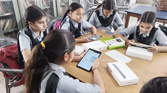 Four female students in school uniforms sit around a wooden table in a library, collaborating while using Samsung tablets and unboxing educational tech kits.