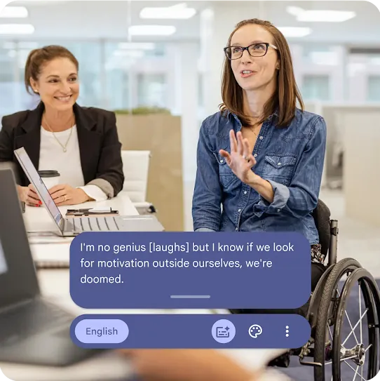 Two women having a conversation in the workplace. One is seated in a wheelchair and wears glasses. The other sits in a chair with a smile.