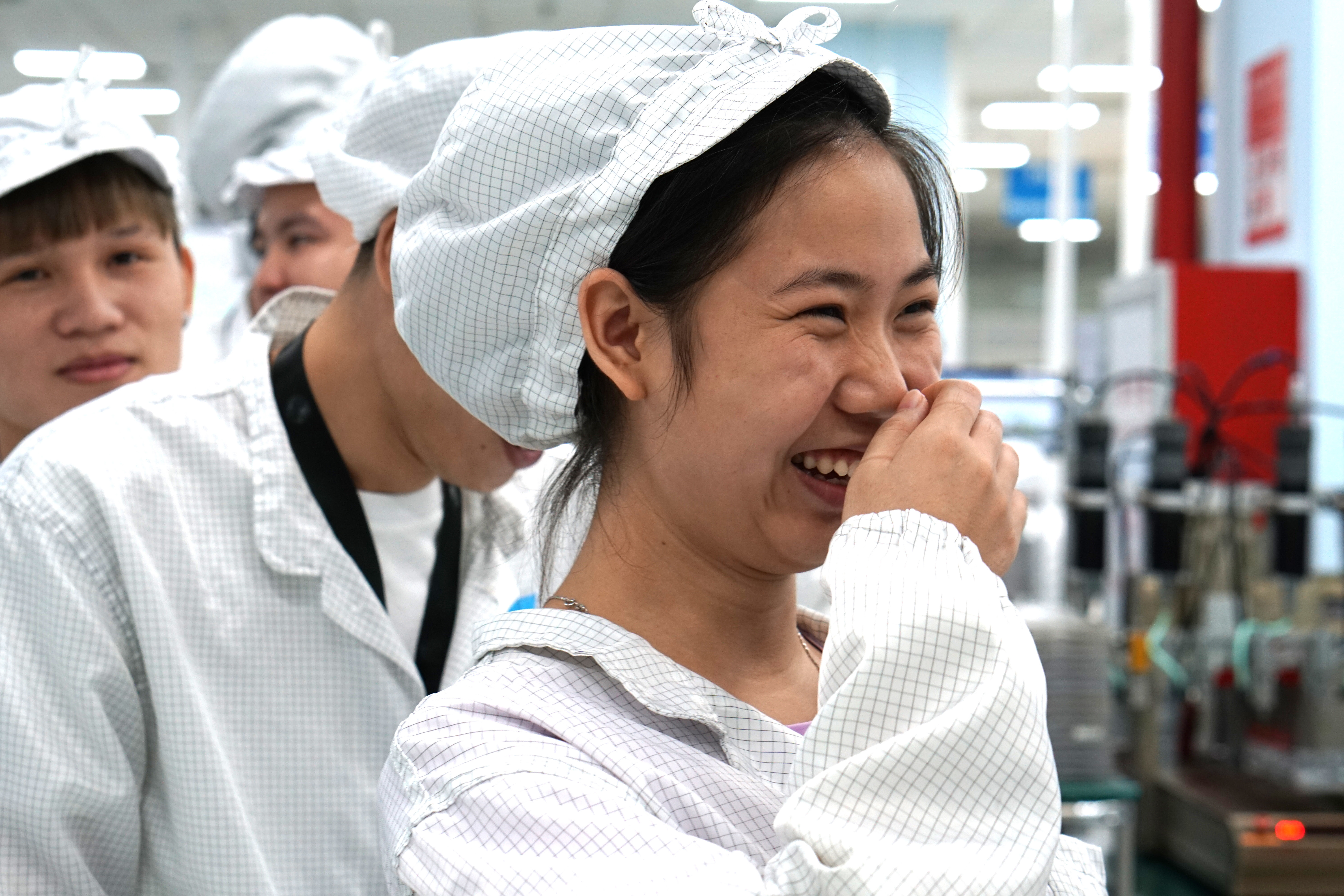 A close up of a factory worker laughing with their hand to their face, wearing a checkered factory uniform.