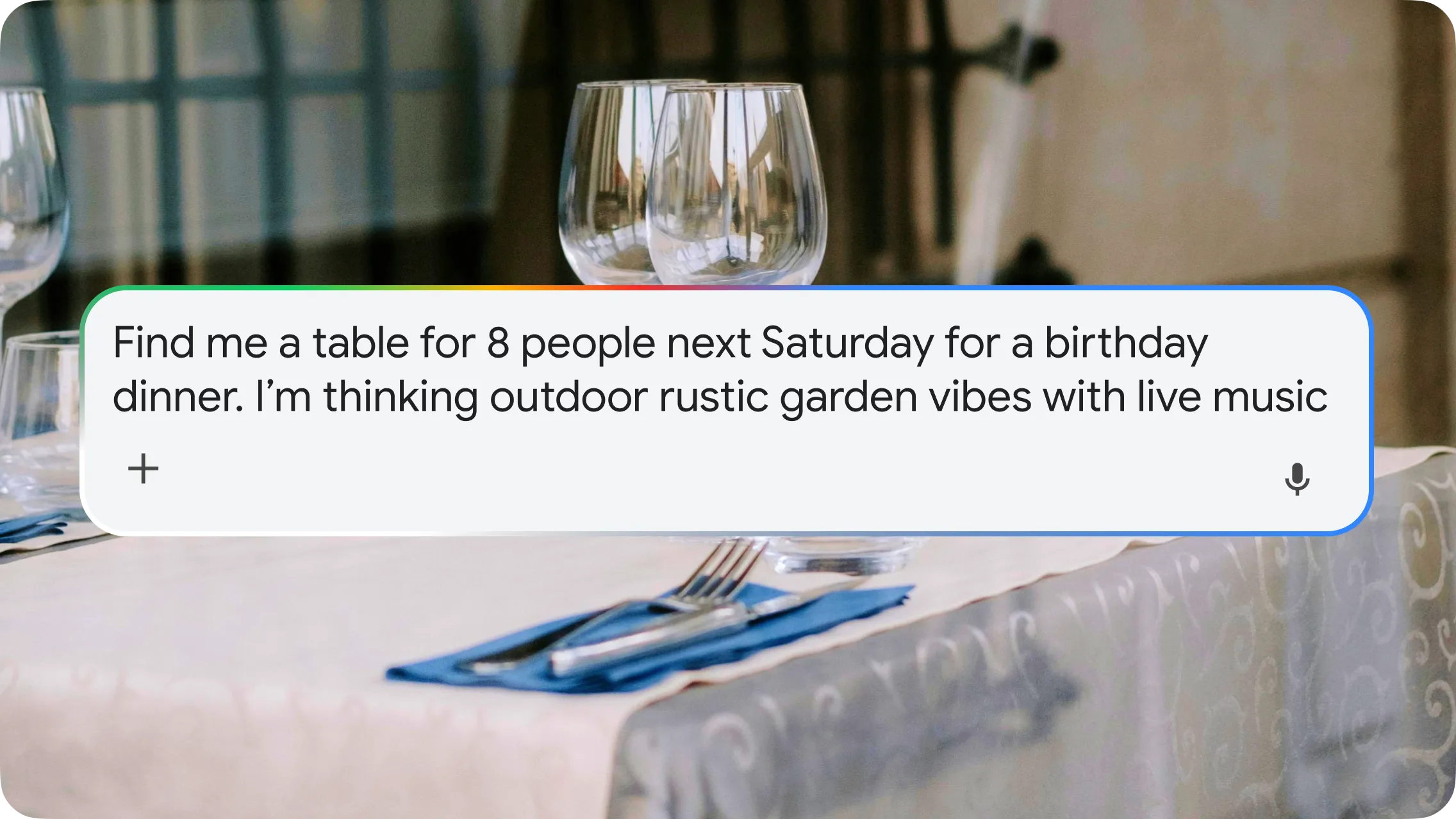 A conversational AI search box overlaid on a blurred background of a set dining table with wine glasses and cutlery. The search box contains the prompt: "Find me a table for 8 people next Saturday for a birthday dinner. I’m thinking outdoor rustic garden vibes with live music." The search bar features a colorful Google-themed glowing top border and a microphone icon.