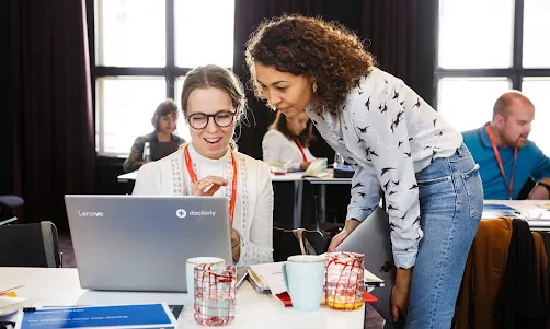 A woman wearing blue top in focus talking to another woman facing away from the camera.