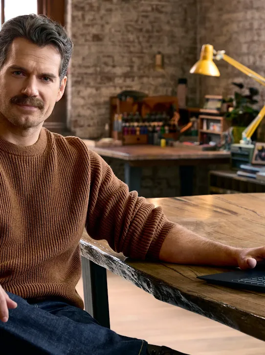 Henry Cavill sitting at a rustic wooden office table with a Googlebook in a sunlit loft.