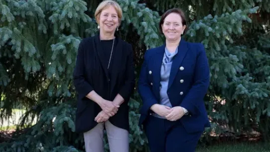 Vivian Schiller, Vice President and Executive Director of Aspen Digital (left), and Heather Adkins, VP of Security Engineering at Google (right), standing together and smiling in front of a large evergreen tree at the 15th Annual Aspen Security Forum.