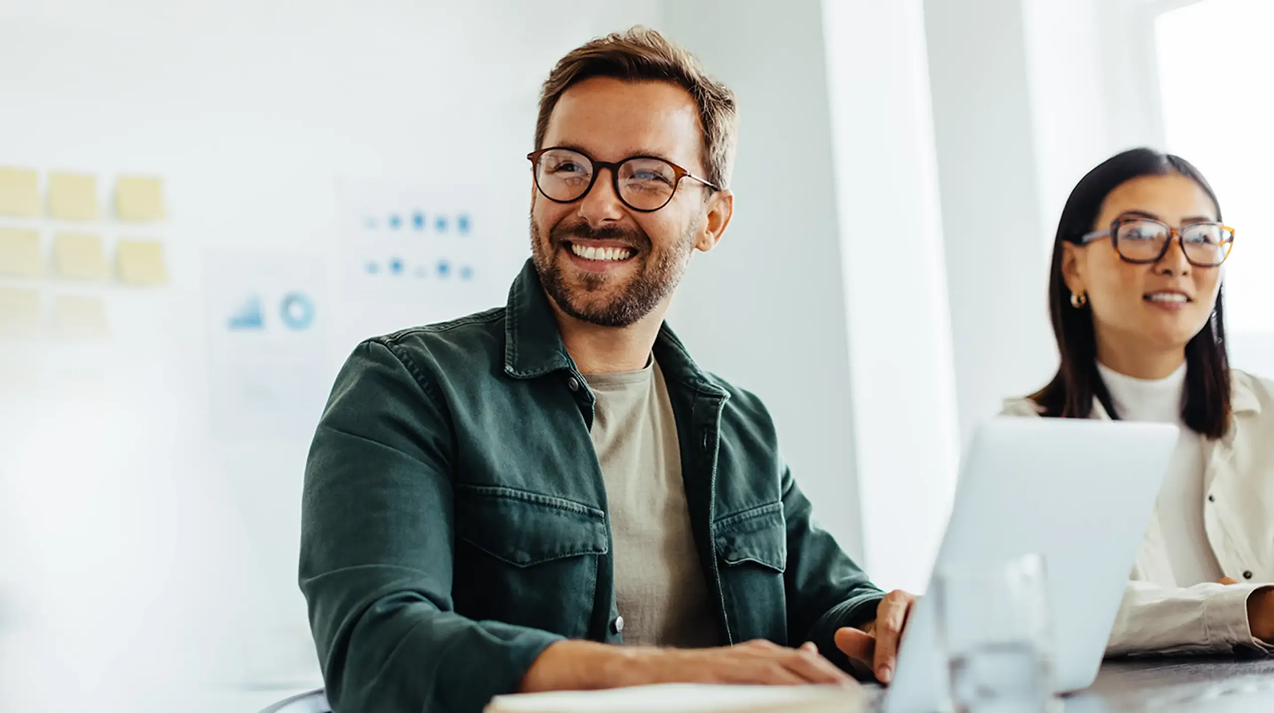A white man with glasses is wearing a beige tee shirt under a green jacket smiling and working on his laptop while sitting in a conference room next to his Asian, female colleague wearing a white turtleneck and glasses.