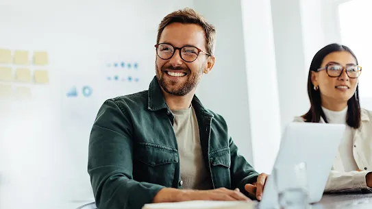 A white man with glasses is wearing a beige tee shirt under a green jacket smiling and working on his laptop while sitting in a conference room next to his Asian, female colleague wearing a white turtleneck and glasses.