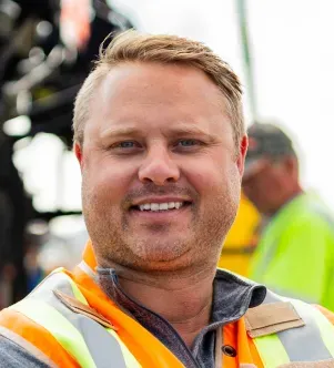 A portrait of Bryce Wuori, co-founder of Pavewise, smiling and wearing an orange high-visibility safety vest on a construction site in North Dakota.