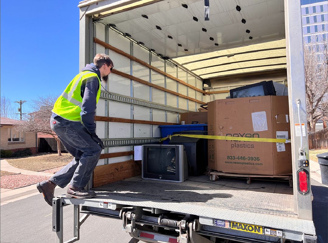 A man in a yellow reflector vests gets into the back of a freight truck filled partially with boxes and equipment