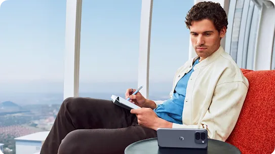 A man sits in a red chair by a large window, writing in a notebook, with a dark grey Google Pixel phone resting on a table in the foreground.