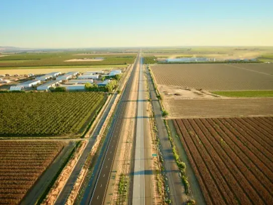 An aerial view looking down a long, straight multi-lane highway stretching to the horizon, flanked on both sides by a patchwork grid of agricultural farm fields, orchards, and rural industrial buildings under a clear, warm sky.