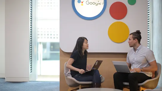 Two colleagues, a woman and a man, sit in comfortable grey armchairs facing each other while engaged in a conversation in a bright, modern office lounge. Both have open laptops resting on their laps, and a small, round wooden coffee table sits between them. Behind them is a large white wall feature adorned with colorful circles in red, yellow, and green, alongside a circular backlit sign featuring the Google Assistant logo. The setting is professional yet relaxed, with large windows in the background allowing for soft, natural light.