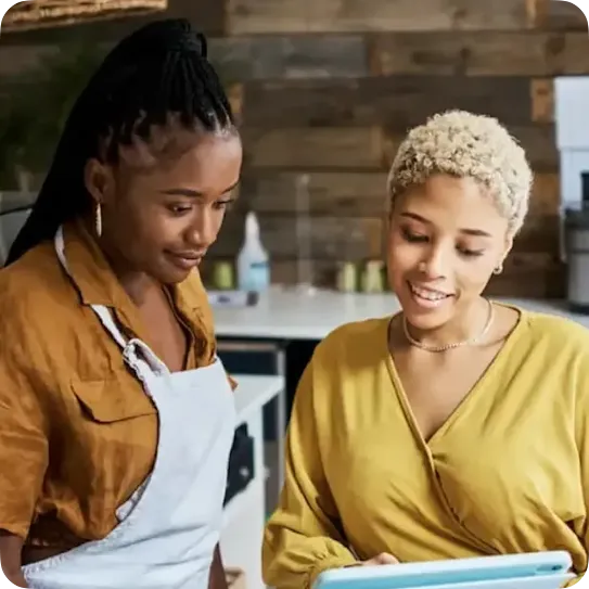 A cafe employee wearing an apron and a manager with short blonde hair looking at a blue tablet screen together behind a counter.