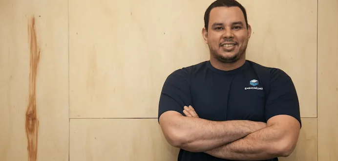 Homem em pé, de braços cruzados, em frente a fundo de madeira.