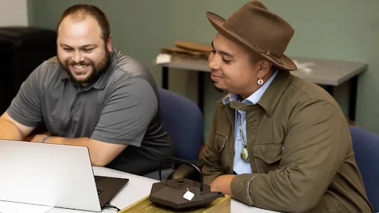 Jake Foreman, a Grow with Google Indigenous Community Digital Coach wearing a brown hat and turquoise jewelry, smiles while leading a digital coaching session at a laptop with a colleague. A cast-iron teapot sits on the table in the foreground.