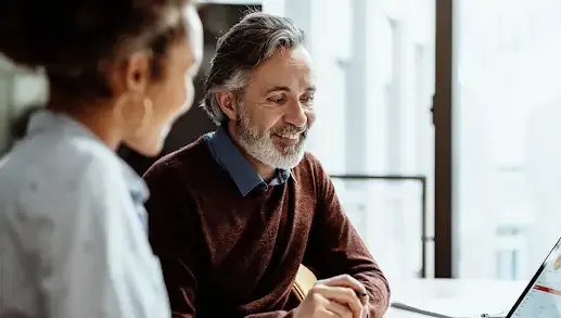 A smiling middle-aged man with grey hair and a beard, wearing a brown sweater over a blue collared shirt, looks attentively at a laptop screen while collaborating with a colleague. The colleague, a woman with dark curly hair tied up, is seen from over her shoulder in the foreground, wearing a light blue shirt. They are seated at a white desk in a bright office setting with windows in the background.