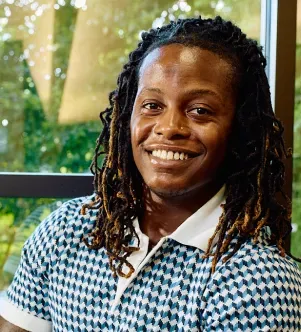 A professional portrait of Jaimeer, a South Carolina graduate student, smiling with long dreadlocks and wearing a geometric patterned polo shirt, featured in a case study about using AI in social work research.