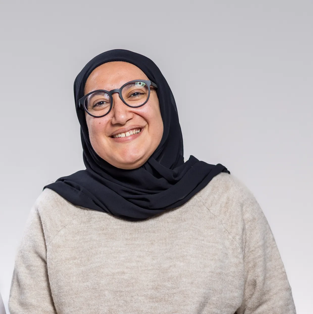 Two smiling women pose side-by-side. One wears a light patterned hijab and white shirt; the other a black hijab and glasses.