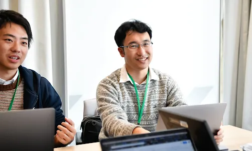 2人の男性がテーブルを挟んで並んで座り、カメラの方を向いている