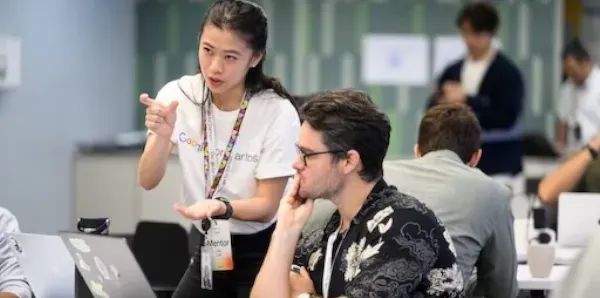 A woman explaining something to a man sitting in front of a laptop in an office setting.