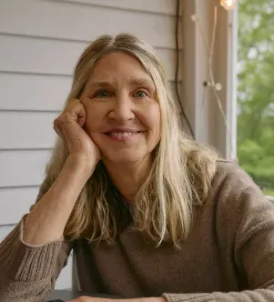 A smiling woman with long blonde hair resting her chin on her hand, wearing a brown sweater in a brightly lit porch area.