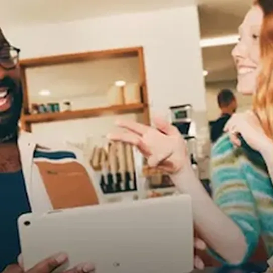 A man wearing glasses and a woman with long auburn hair are interacting in a cafe or casual workplace setting. The man is showing something on a tablet to the woman, who is smiling and looking at the man.