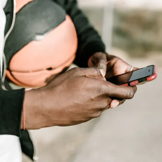 Close-up of a person's hands using a smartphone with a red case while holding a basketball under their arm.