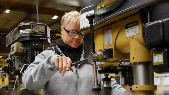 A woman wearing safety glasses and a grey hoodie operates a heavy-duty yellow Powermatic drill press in a well-lit manufacturing facility.