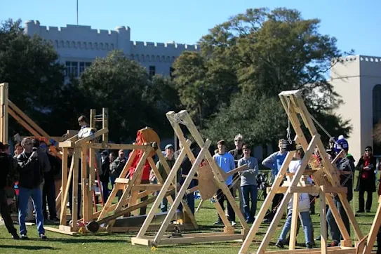 People gathered outdoors around wooden trebuchets and catapult structures set up on a grassy field