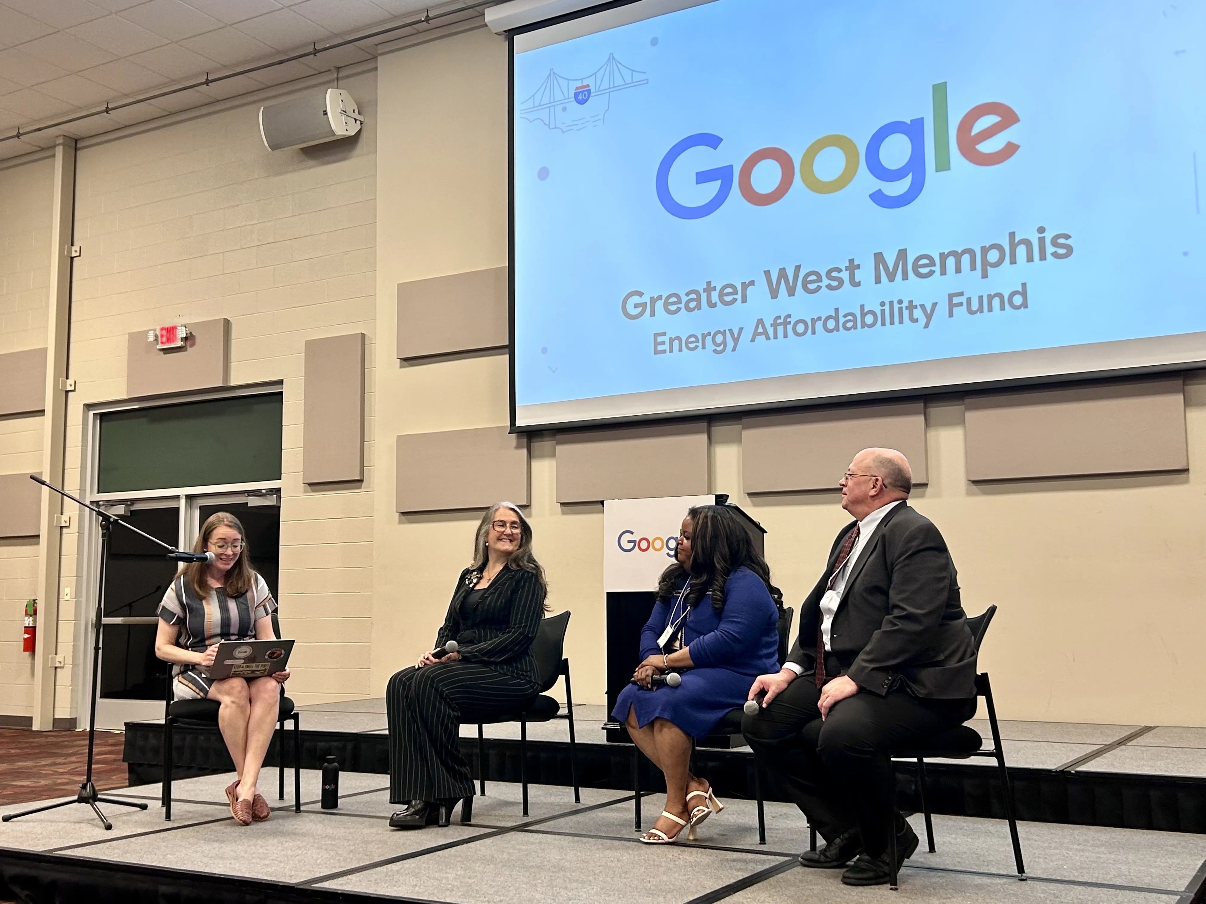 A panel of four people sitting on a stage during a presentation. A large projector screen in the background displays the Google logo and the text "Greater West Memphis Energy Affordability Fund." One woman on the left is speaking into a microphone while looking at a laptop, while the other three participants listen and smile.