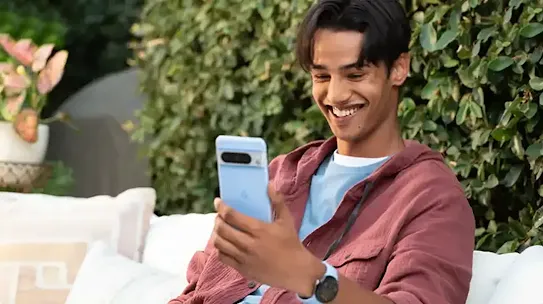 A man sitting on a patio outdoors while looking at a smartphone and smiling.