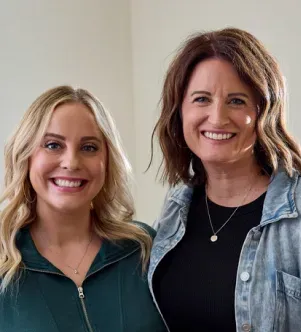Two smiling women standing together; the woman on the left has blonde hair and a green top, and the woman on the right has brown hair and a denim jacket over a black shirt.