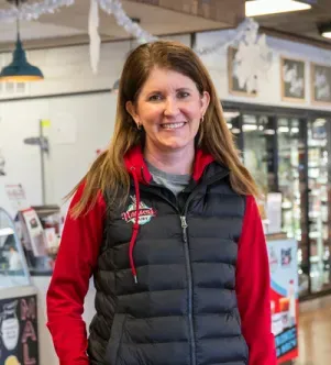 A smiling woman with long brown hair wearing a red hoodie and a black puffer vest with a "Hansen's Dairy" logo, standing inside a bright dairy store or cafe.