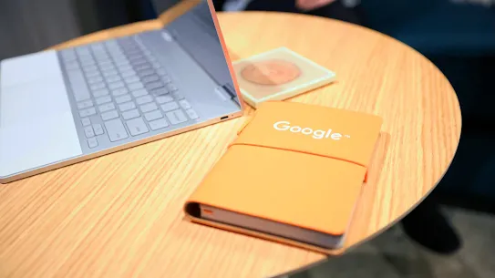 On a table is an orange notebook with the logo Google on it. Next to the notebook is a computer. 