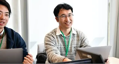 Two men, one wearing glasses, are having a conversation and smiling while working on laptops.