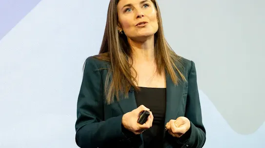 A professional woman with long brown hair, wearing a dark teal blazer, speaking on stage at the "Growing Up in the Digital Age" summit. She is holding a slide clicker and gesturing during her presentation. In the background, a white screen displays the partial word "EMPOWER" in gold lettering.