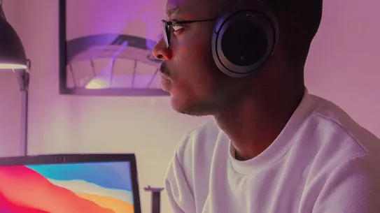 A young Black man wearing glasses and black over-ear headphones looking focused while working in front of a laptop under purple ambient light.