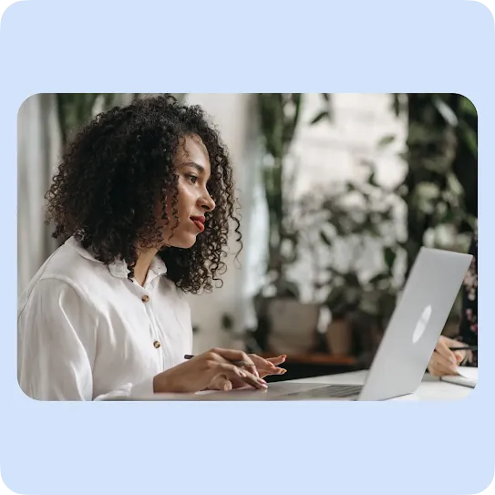 A woman with curly dark hair and a white button-down shirt looks intently at her laptop screen while typing at a white desk. In the blurred background, green houseplants are visible, creating a bright and modern remote work or office environment.