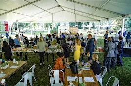 A high-angle wide shot of an outdoor event held under a large white tent on a grassy lawn. In the background, a "Safer with Google" panel is being filmed, while in the foreground, attendees gather around buffet tables and sit at long wooden picnic-style tables. The setting is lush with green trees and park benches visible outside the tent.