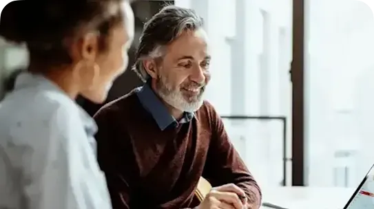 A smiling middle-aged man with grey hair and a beard, wearing a brown sweater over a blue collared shirt, looks attentively at a laptop screen while collaborating with a colleague. The colleague, a woman with dark curly hair tied up, is seen from over her shoulder in the foreground, wearing a light blue shirt. They are seated at a white desk in a bright office setting with windows in the background.