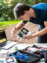 A person in a blue polo shirt examines an open electronic sensor box outdoors, with a laptop and tools on a table in the foreground. 