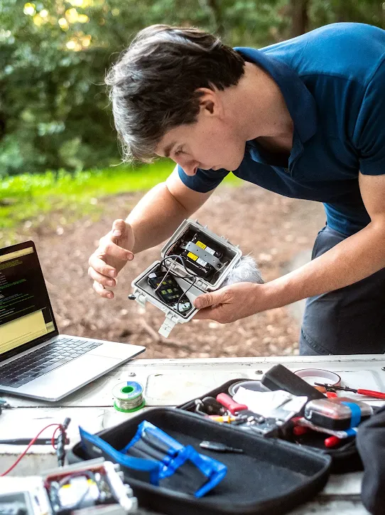 A person in a blue polo shirt examines an open electronic sensor box outdoors, with a laptop and tools on a table in the foreground. 