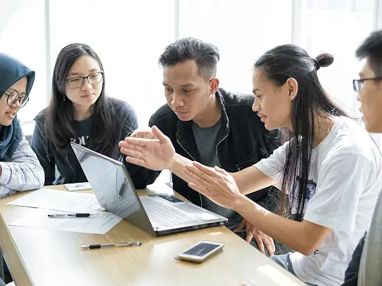 Five young colleagues engaged in a discussion, with one person pointing at a laptop screen during a collaborative session.