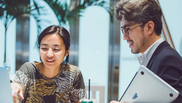 A woman showing something to a man on laptop