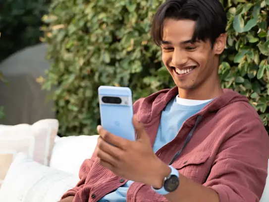 A man sits outdoors while looking at a smartphone and smiling.