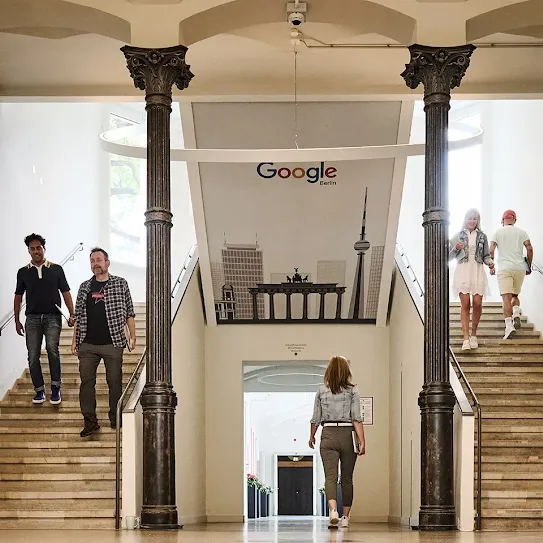 Eine Gruppe von vier Personen arbeitet in einem modernen, hellen Büro zusammen. Drei lachen vor einem Laptop, während eine Frau winkt; eine weitere Person arbeitet im Hintergrund.