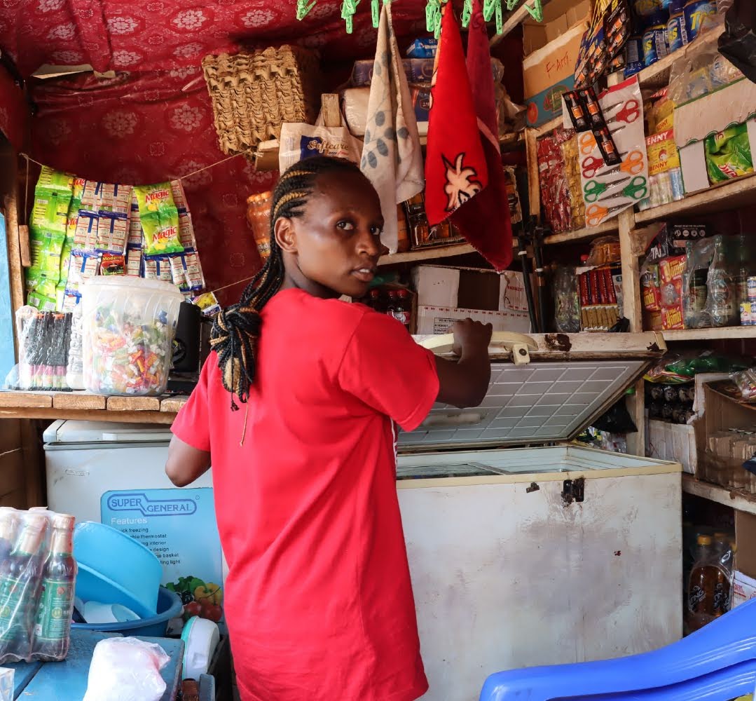 A vendor wearing a red shirt stands in a shop propping up the lid of a cooler, with walls lined with items like snacks, scissors and supplies